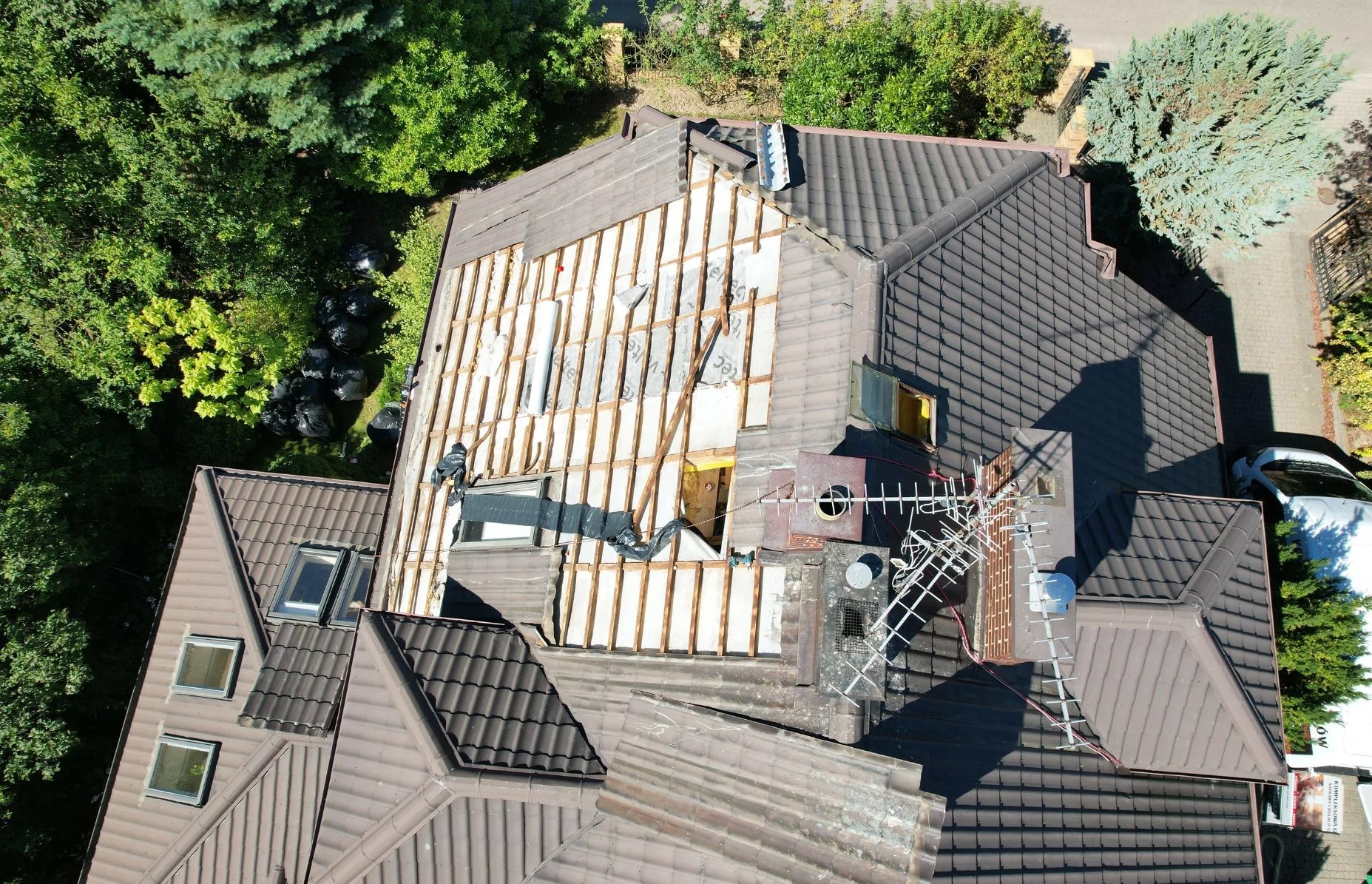 Demontaż starego pokrycia dachowego z widoczną konstrukcją i ociepleniem. Prace dekarskie w toku, widok z drona na brązowy dach i czarne worki na odpady.