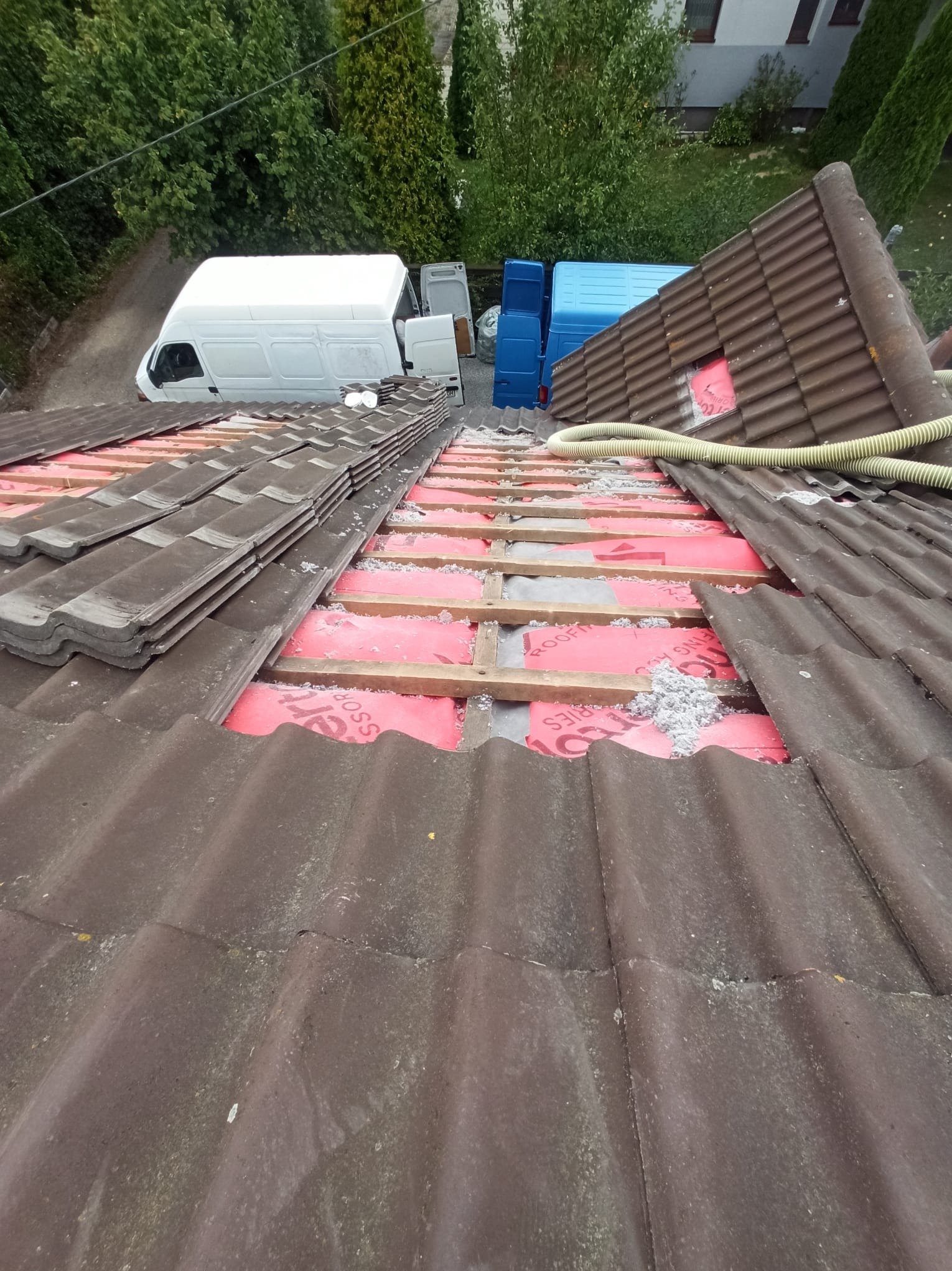 Montaż izolacji na dachu z częściowo zdjętą dachówką. Widoczne łaty, izolacja w kolorze czerwonym i biała wełna. W tle zaparkowane busy.
