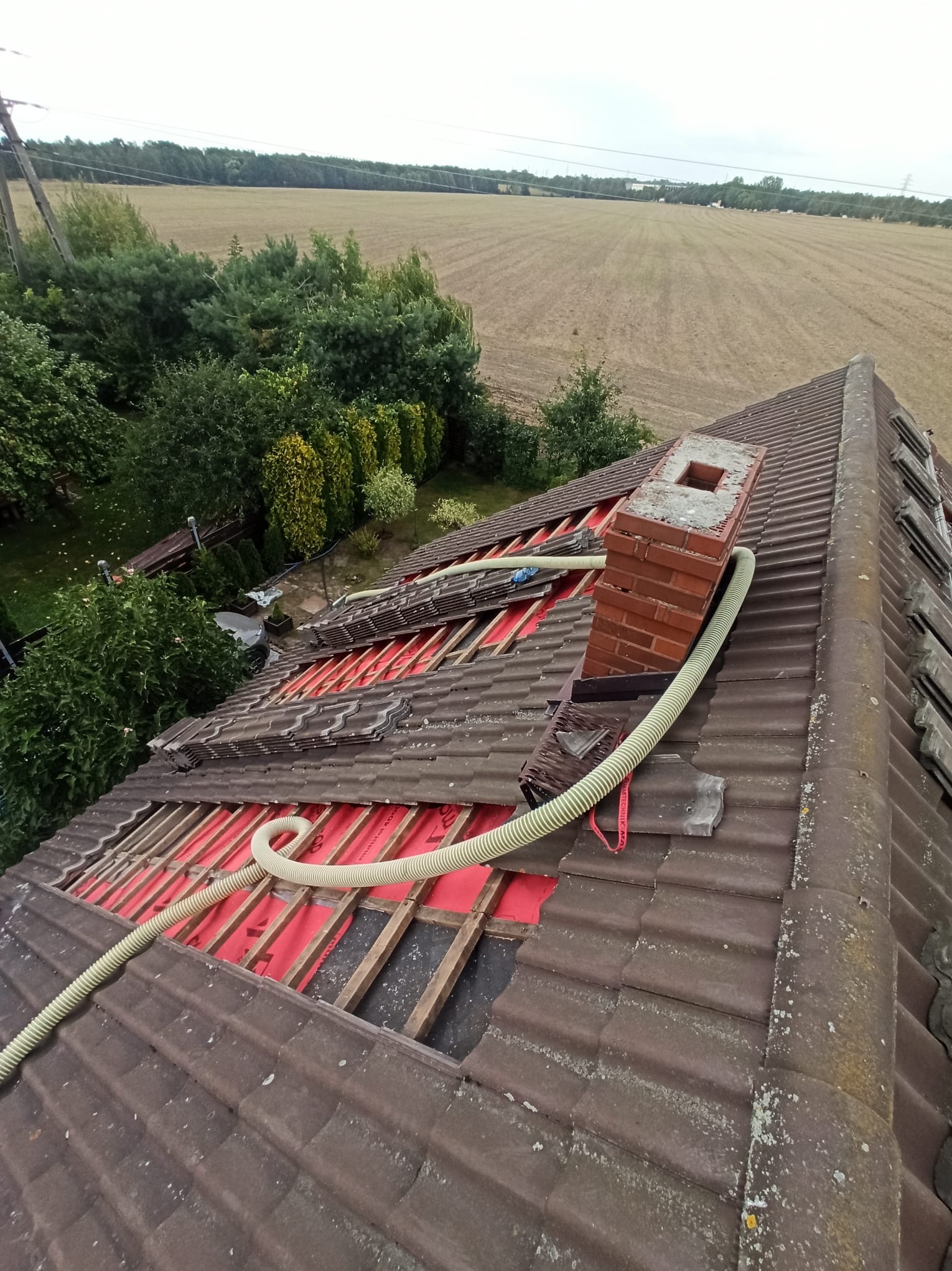 Demontaż dachówki wokół komina, widoczne czerwone membrany i rury wentylacyjne. W tle zielona roślinność i pole uprawne pod pochmurnym niebem.