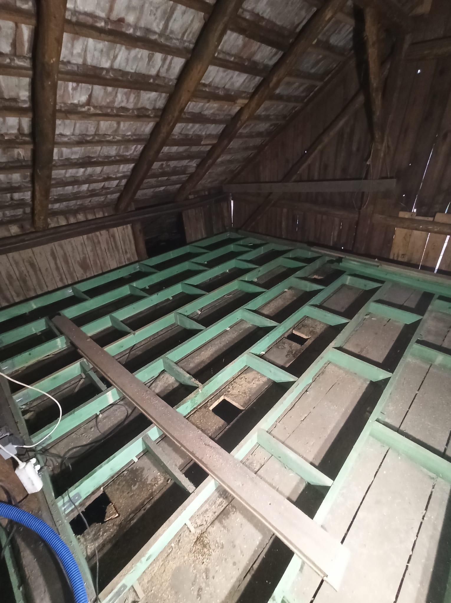 Konstrukcja poddasza z drewnianymi belkami w zielonym kolorze, przygotowana do dalszych prac. Widoczne stare deski i elementy instalacji.