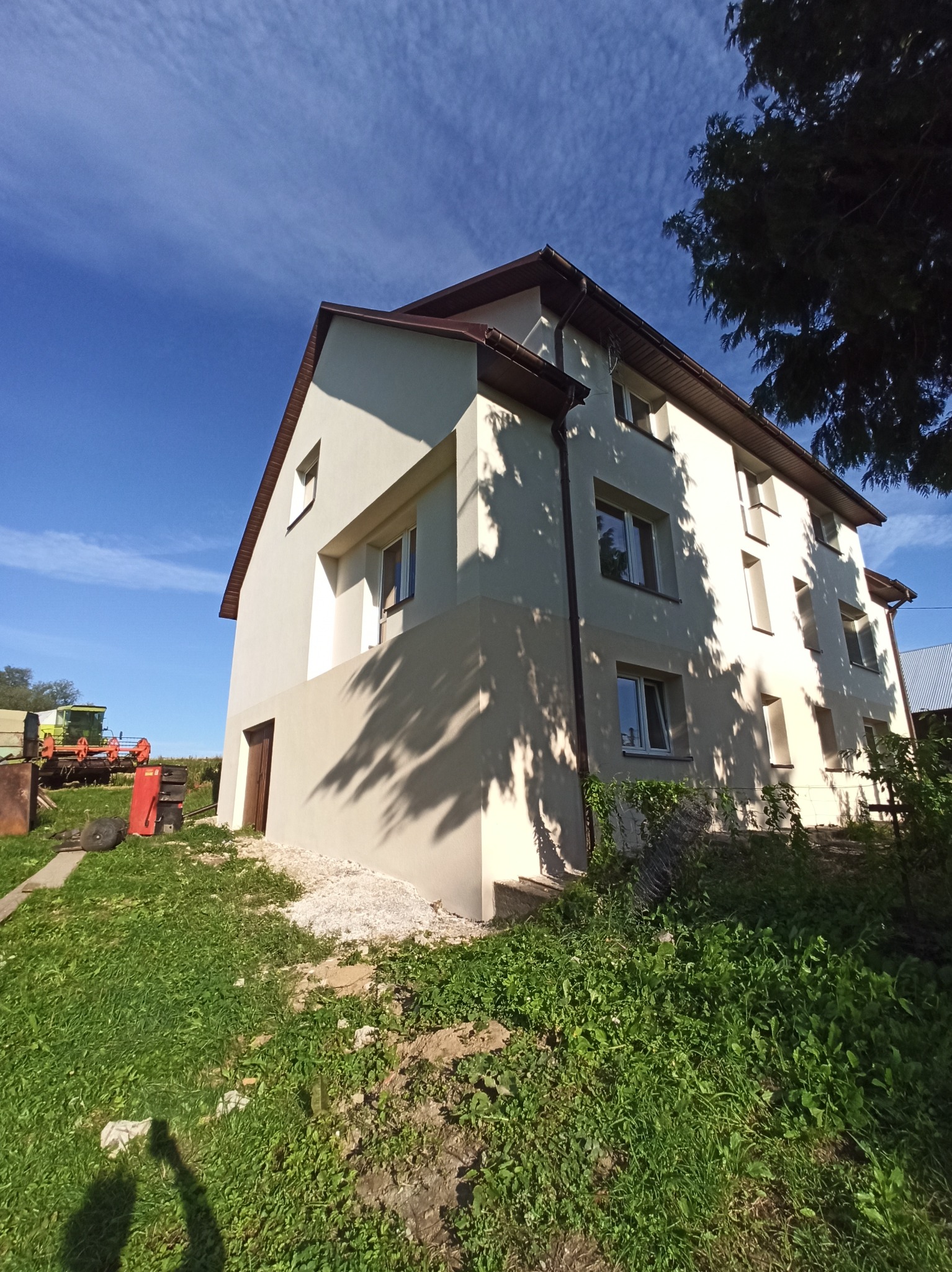 Dwupiętrowy dom po renowacji elewacji w słoneczny dzień. Jasna fasada, brązowy dach i okna. Na trawniku sprzęt rolniczy. Błękitne niebo z chmurami w tle.