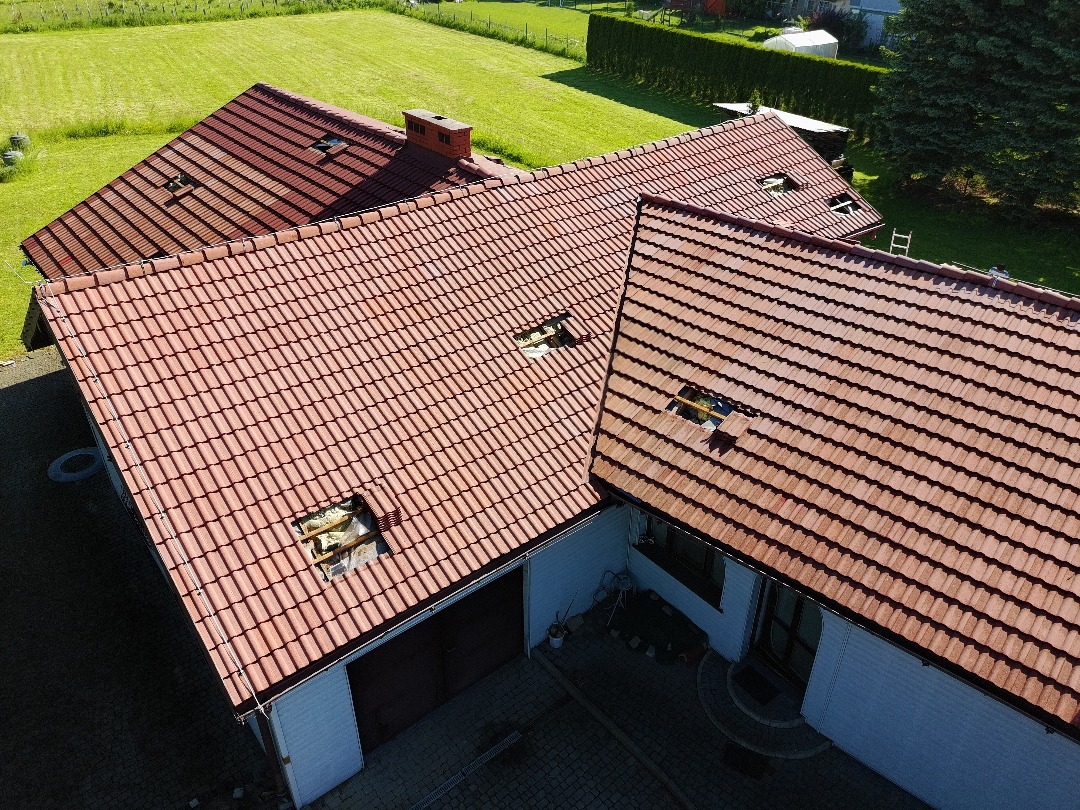 Widok z góry na dom z czerwoną dachówką, z widocznymi fragmentami ocieplenia w miejscach uszkodzeń. Zielony trawnik w tle, słoneczny dzień.