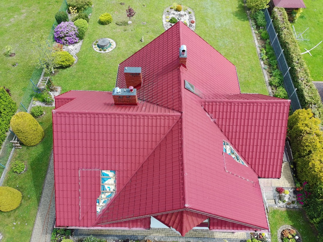 Czerwony dach domu z blachodachówki, widok z góry na tle zielonego ogrodu. Widoczne kominy i okno dachowe. Ujęcie z drona.