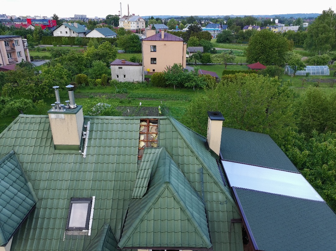 Zielony dach z kominem i oknem dachowym, widok z góry na tle zabudowy miejskiej i zieleni. Fragment dachu z uszkodzeniami. Nietypowe ujęcie z góry.