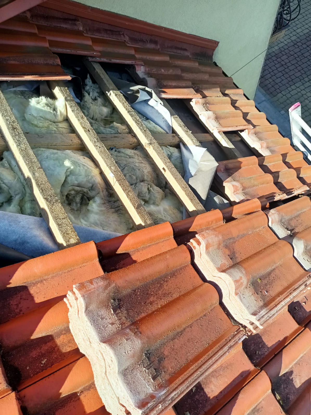 Odsłonięty dach z widoczną izolacją z wełny mineralnej między krokwiami, częściowo pokryty czerwoną dachówką. Fragment dachu w trakcie prac remontowych.