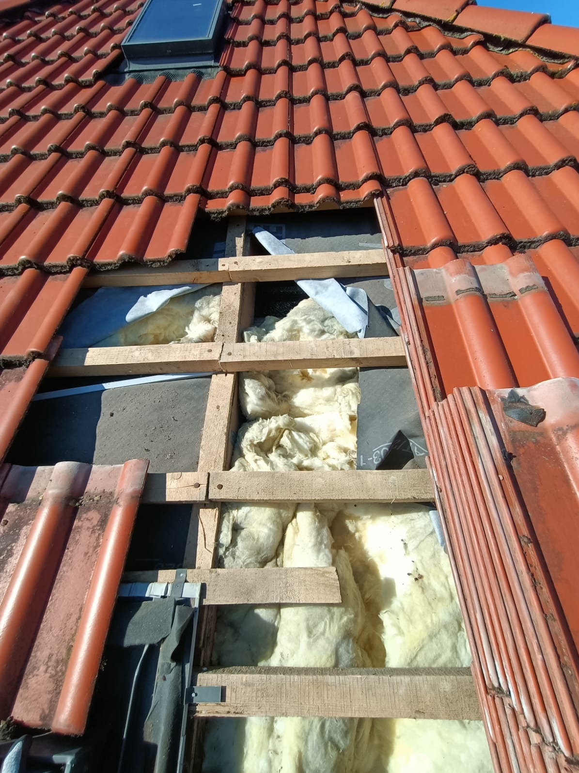 Częściowo zdjęte dachówki ukazują drewnianą konstrukcję i izolację z wełny mineralnej. Widoczne okno dachowe i fragmenty czerwonej dachówki.