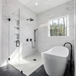 Nowoczesna łazienka z kabiną prysznicową typu walk-in, białą wanną wolnostojącą i czarną armaturą na tle marmurowych ścian i ciemnej podłogi.