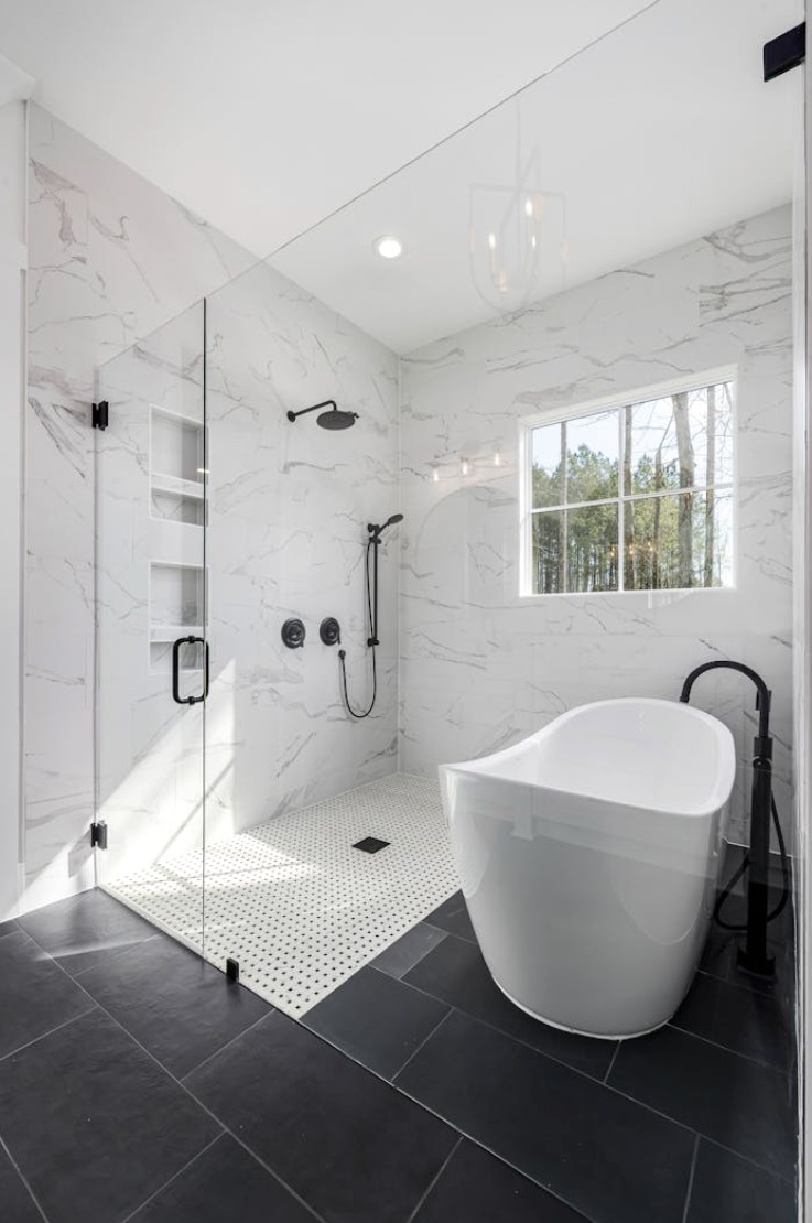 Nowoczesna łazienka z kabiną prysznicową typu walk-in, białą wanną wolnostojącą i czarną armaturą na tle marmurowych ścian i ciemnej podłogi.