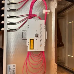 Srebrna skrzynka z panelami światłowodowymi i r&oacute;żowymi kablami, z zamontowanym modułem laserowym klasy 1, ukazująca precyzyjne połączenia i uporządkowany układ.