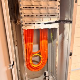 Klimczyk-Elektro - Szafa rack z patch panelem i uporządkowanymi kablami światłowodowymi w kolorze pomarańczowym. Widoczna listwa zasilająca i oświetlenie robocze.