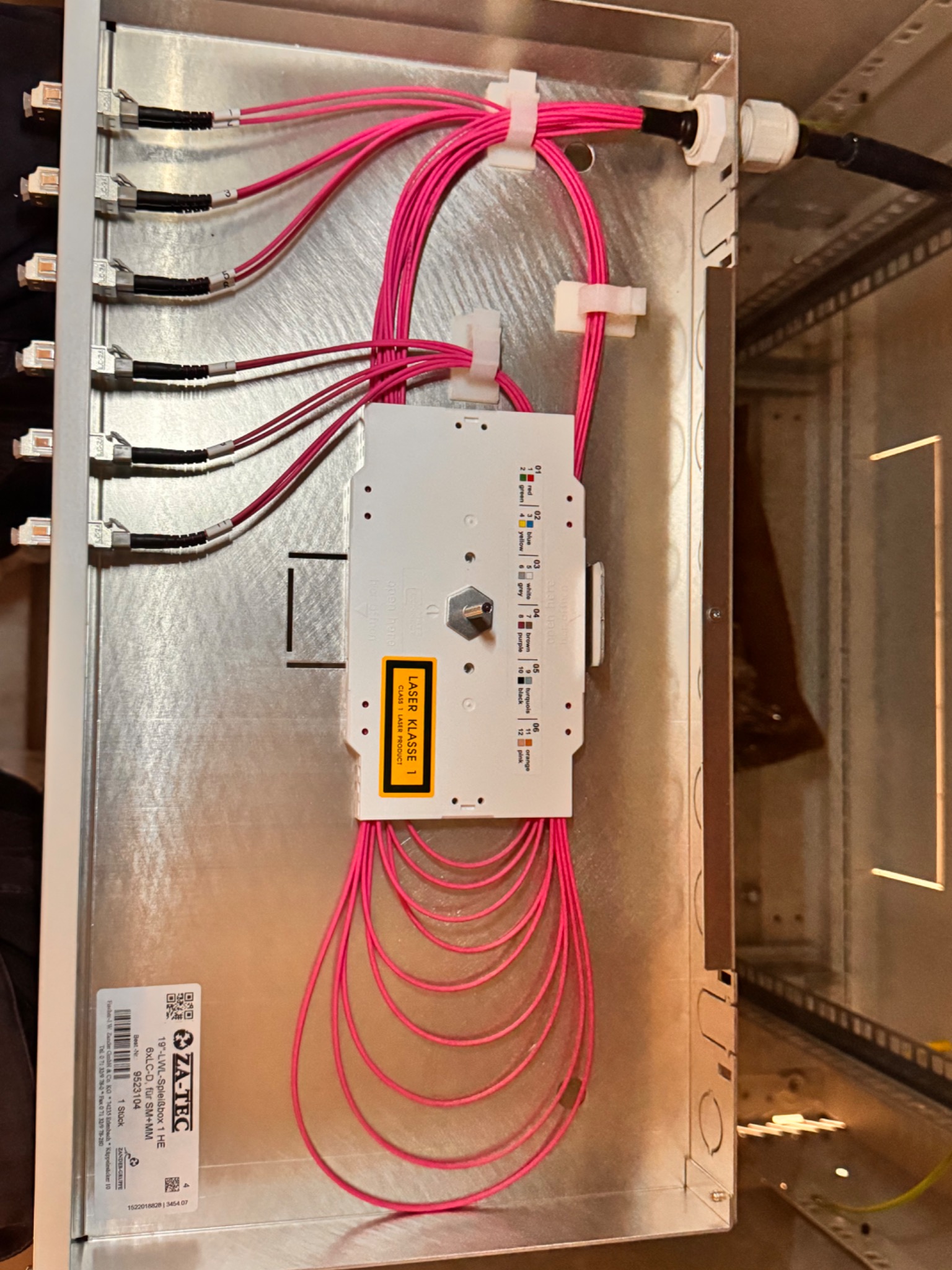 Srebrna skrzynka z panelami światłowodowymi i różowymi kablami, z zamontowanym modułem laserowym klasy 1, ukazująca precyzyjne połączenia i uporządkowany układ.