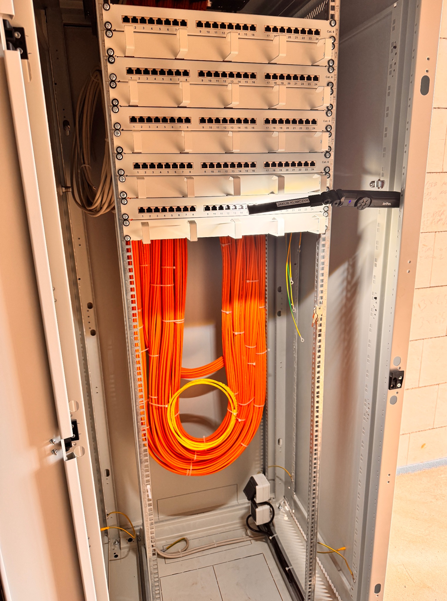 Szafa rack z patch panelem i uporządkowanymi kablami światłowodowymi w kolorze pomarańczowym. Widoczna listwa zasilająca i oświetlenie robocze.