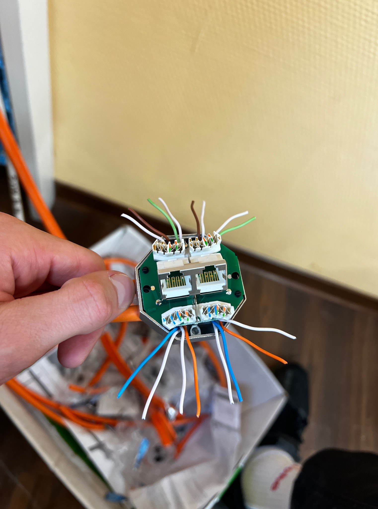 Podłączenie kabli do modułu sieciowego. Widoczna płytka z gniazdami i kolorowe przewody. Pomarańczowe kable w tle. Praca w toku, etap łączenia przewodów.