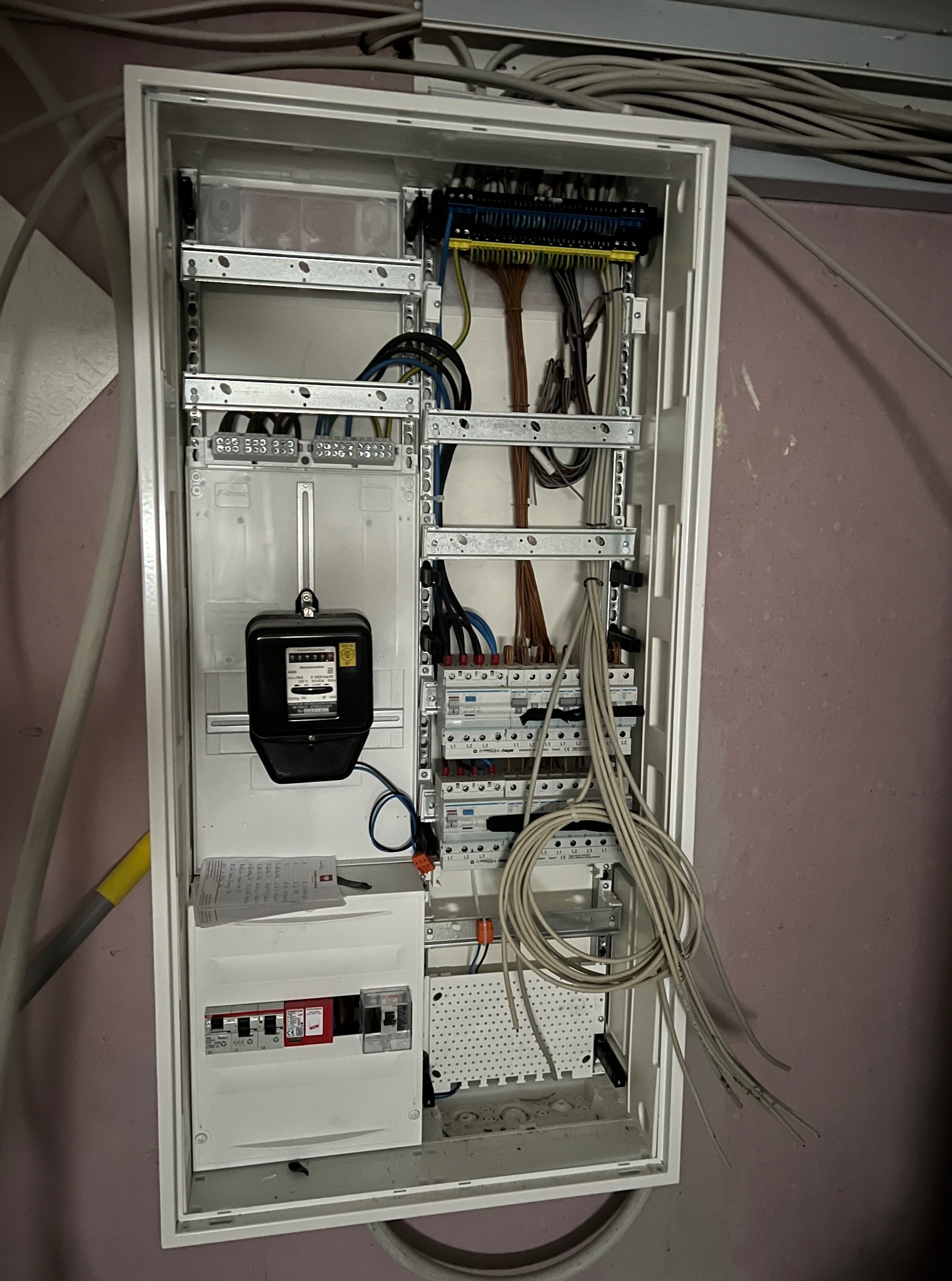 Otwarta, biała skrzynka elektryczna z licznikiem, bezpiecznikami i przewodami. Widoczne elementy instalacji elektrycznej na tle różowej ściany i rur.