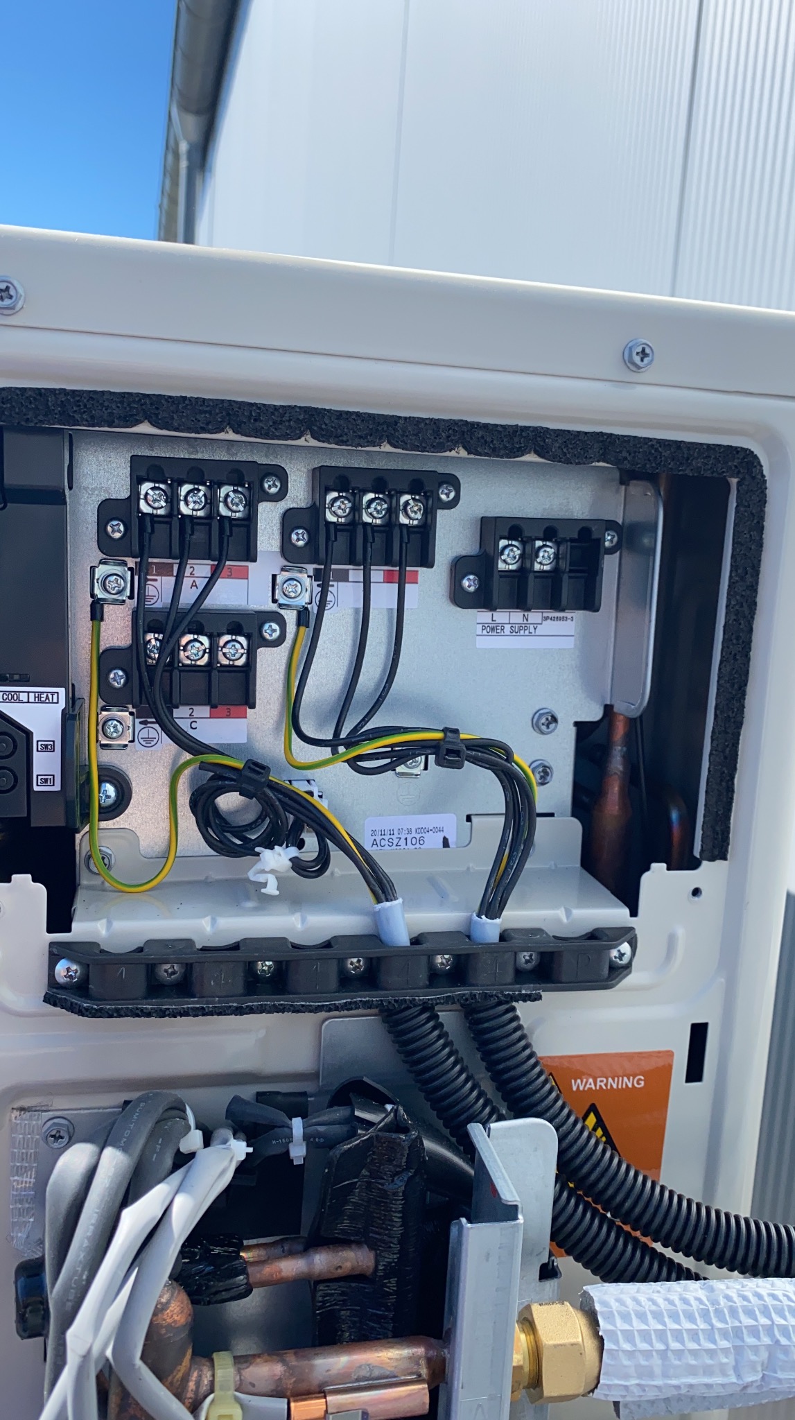 Wnętrze jednostki zewnętrznej klimatyzacji z widocznymi podłączeniami elektrycznymi i kablami. Detaliczne ujęcie na złącza i przewody w obudowie urządzenia.