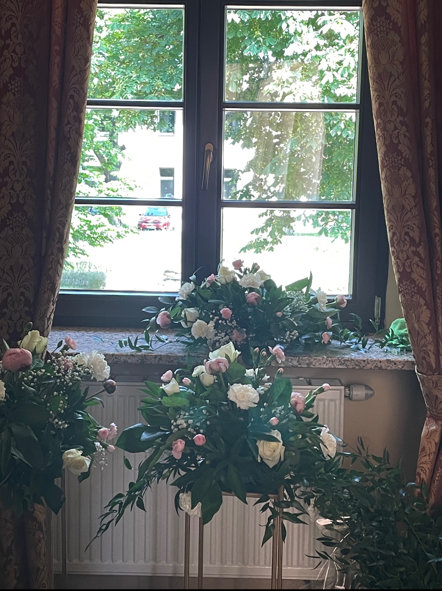 Dekoracja kwiatowa z białymi i różowymi kwiatami na parapecie okna z widokiem na zieleń. W tle zasłony w kwiatowy wzór i grzejnik.