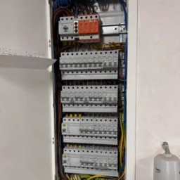 Otwarta skrzynka rozdzielcza z zainstalowanymi bezpiecznikami, przewodami i listwami zaciskowymi. Widoczne elementy instalacji elektrycznej wewnątrz obudowy.