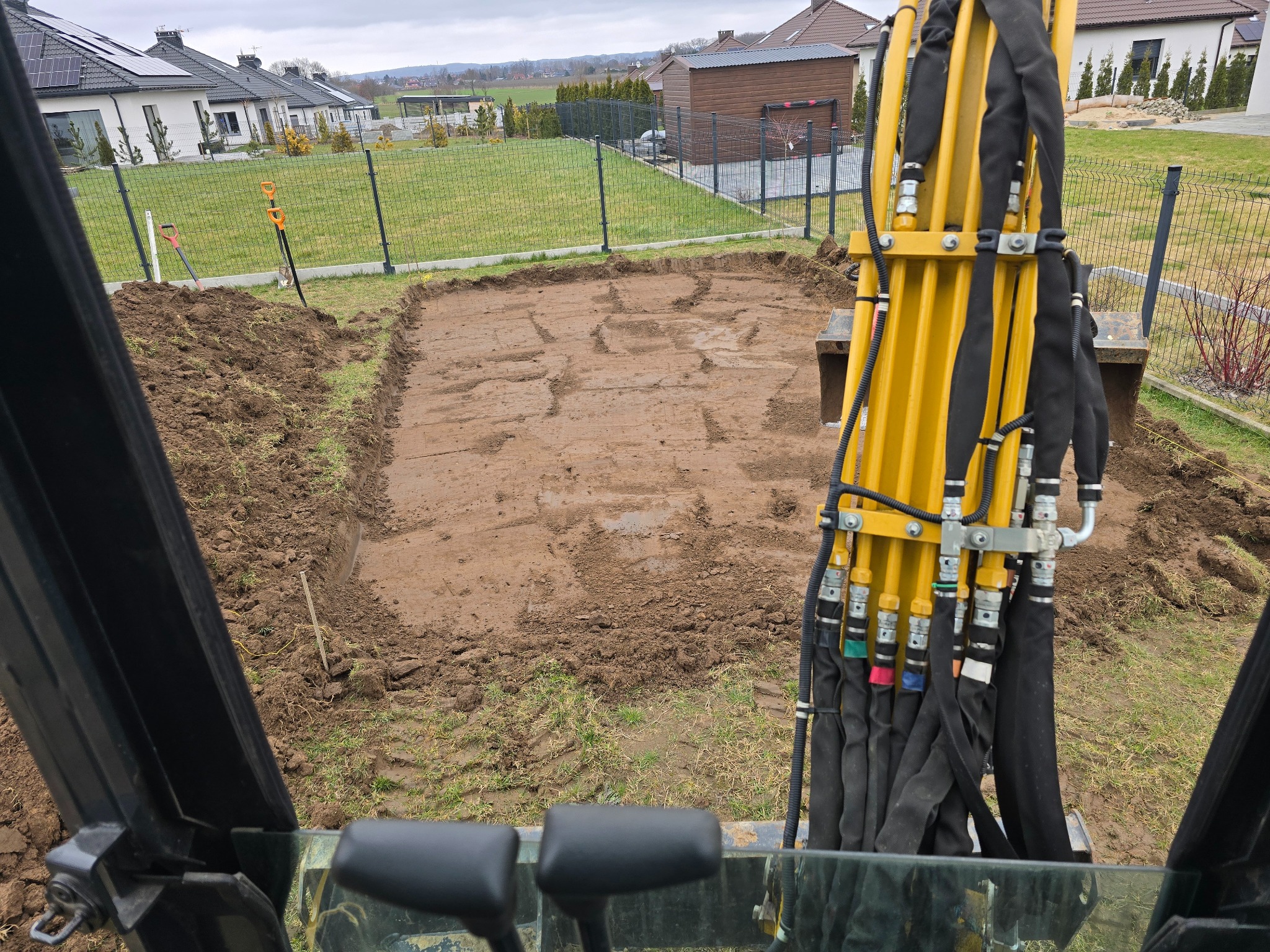 Widok z kabiny koparki na wykop pod fundamenty w ogrodzie, narzędzia w tle. Specyficzne ułożenie przewodów hydraulicznych koparki.