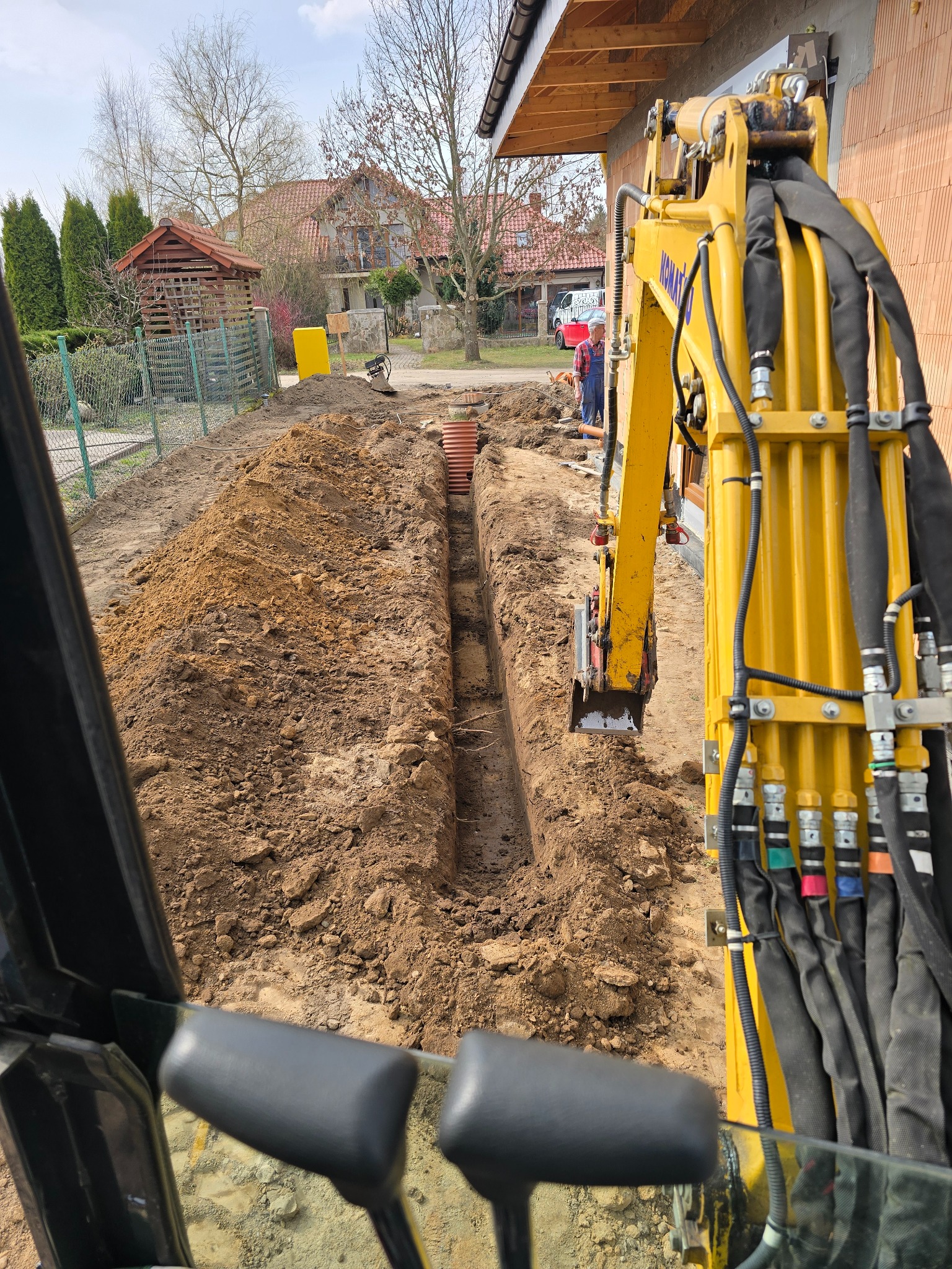 Żółta koparka Komatsu w trakcie prac ziemnych, widok z kabiny. Wykop pod rury na tle zabudowy jednorodzinnej.