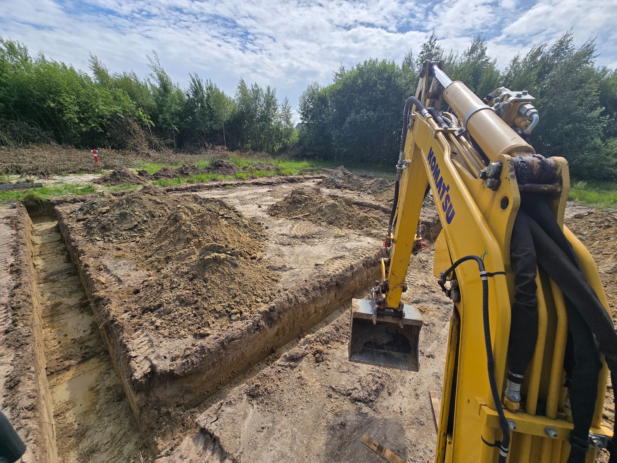 Żółta koparka Komatsu w trakcie prac ziemnych, wykop pod fundamenty na działce z drzewami w tle. Widoczne świeżo wykopane rowy i hałdy ziemi.