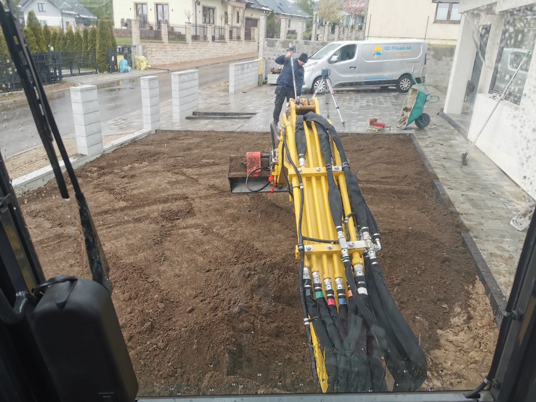 Żółta minikoparka wyrównuje teren pod kostkę na posesji w Skwierzynce. Widok z kabiny operatora, w tle pracownik z niwelatorem i firmowy bus.