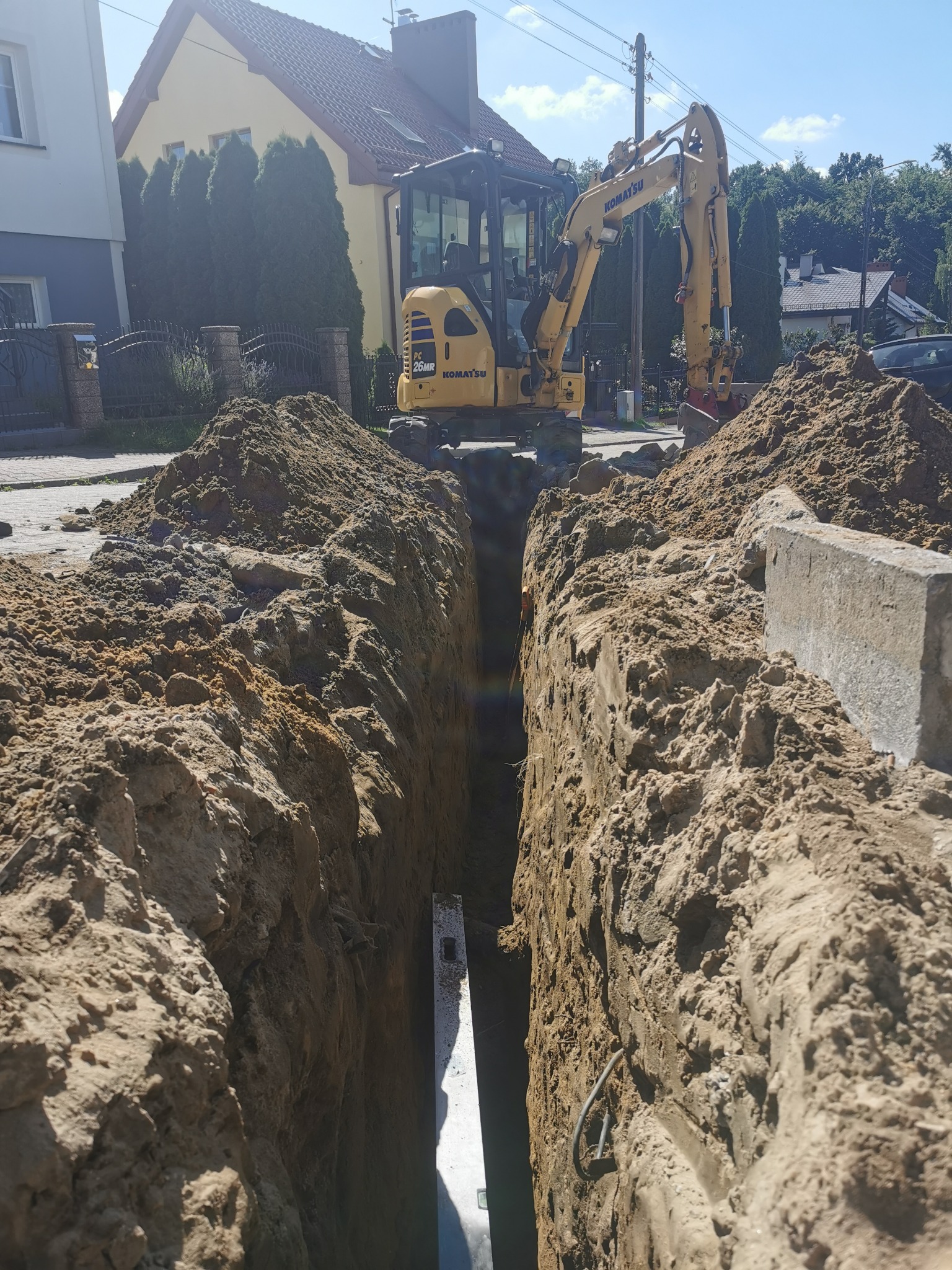 Wąski wykop w ziemi z widoczną żółtą koparką Komatsu w tle, częściowo zasłoniętą przez usypaną ziemię. Wykop zabezpieczony stalowymi elementami.