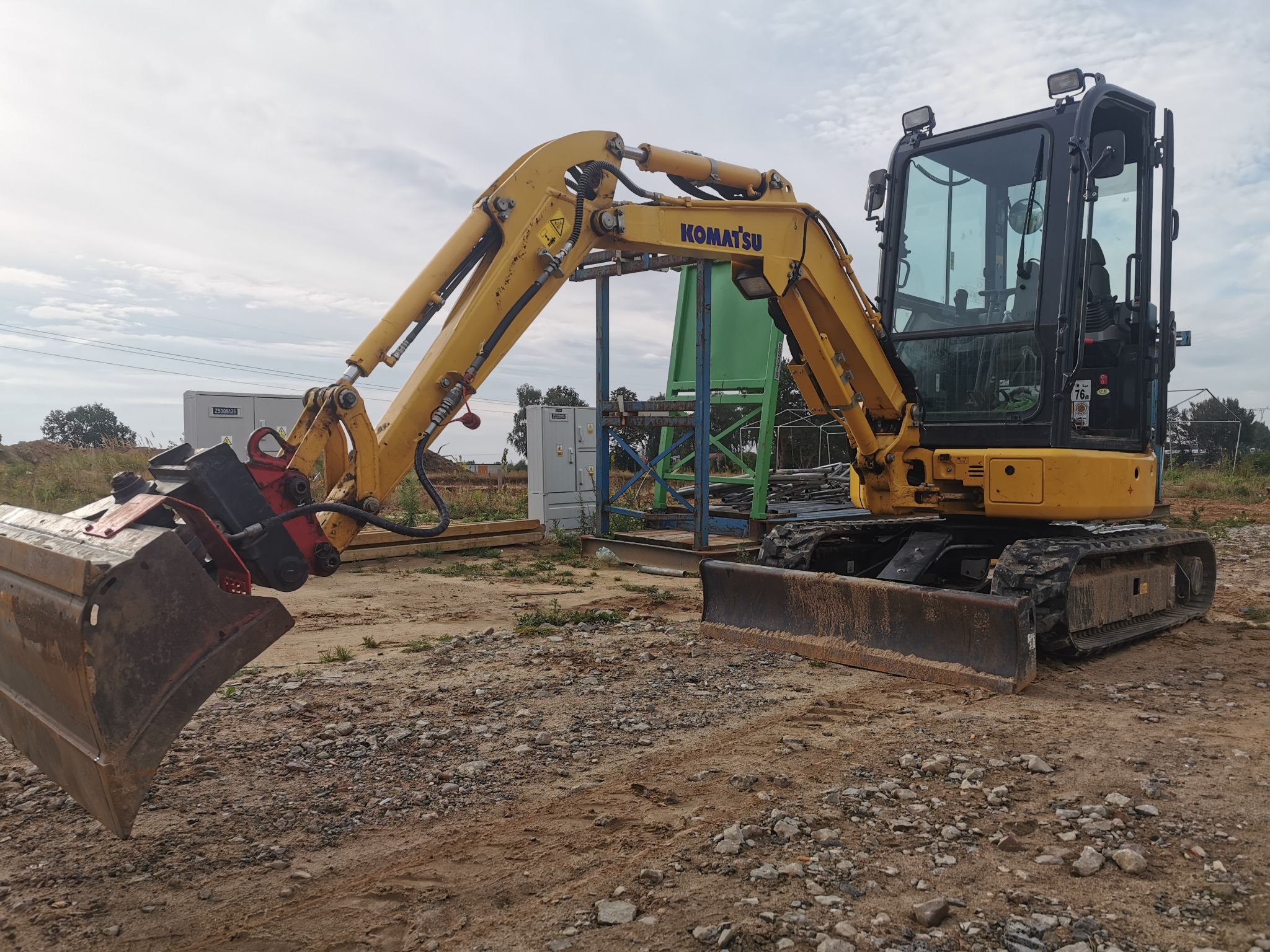Żółta koparka Komatsu na żwirowym terenie. Widoczny lemiesz i łyżka koparki, a w tle skrzynka elektryczna i konstrukcja stalowa. Pochmurne niebo.