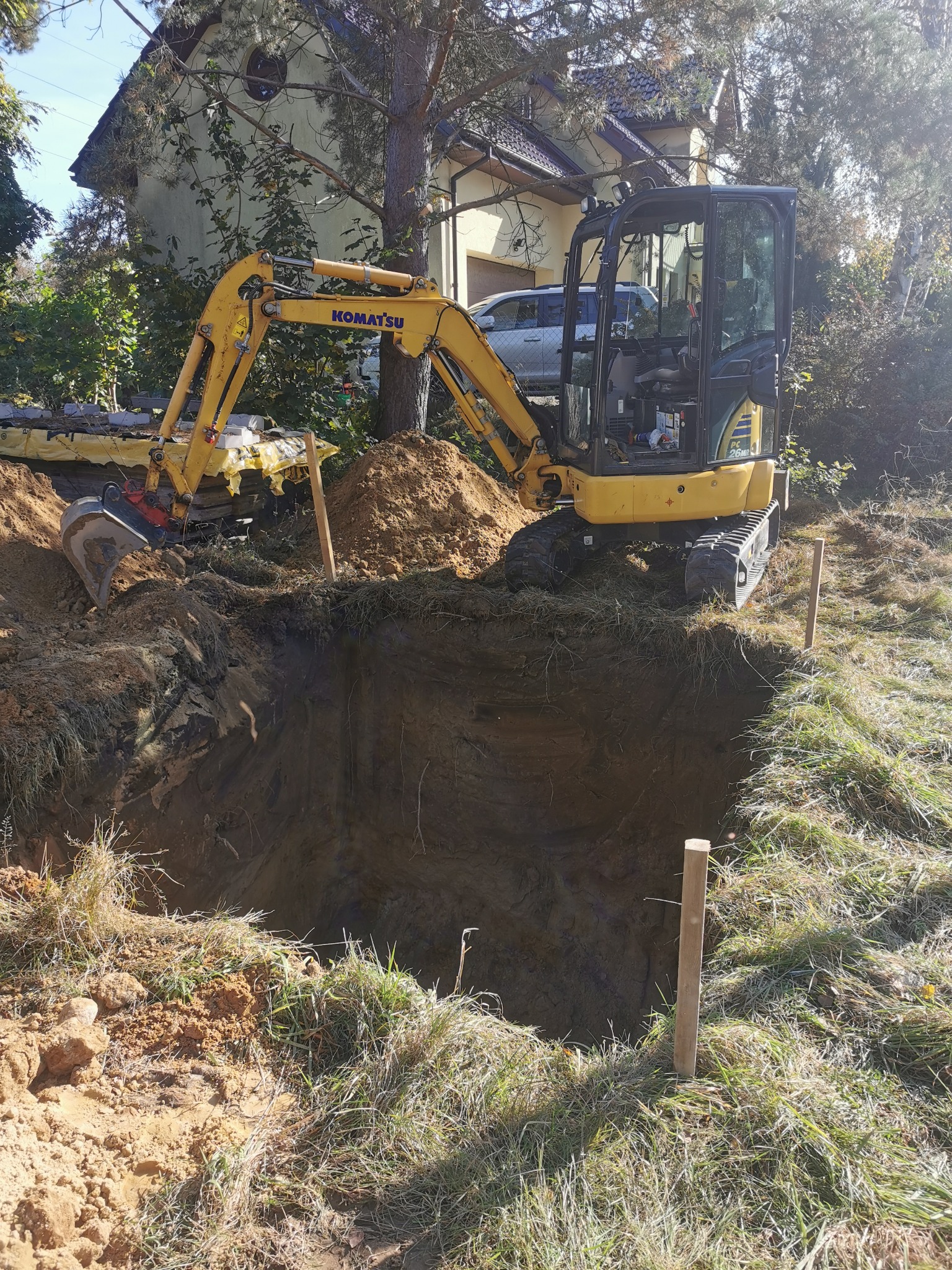 Żółta mini koparka Komatsu PC26MR stoi obok głębokiego wykopu, drewniane paliki wyznaczają jego granice, w tle dom i samochód.