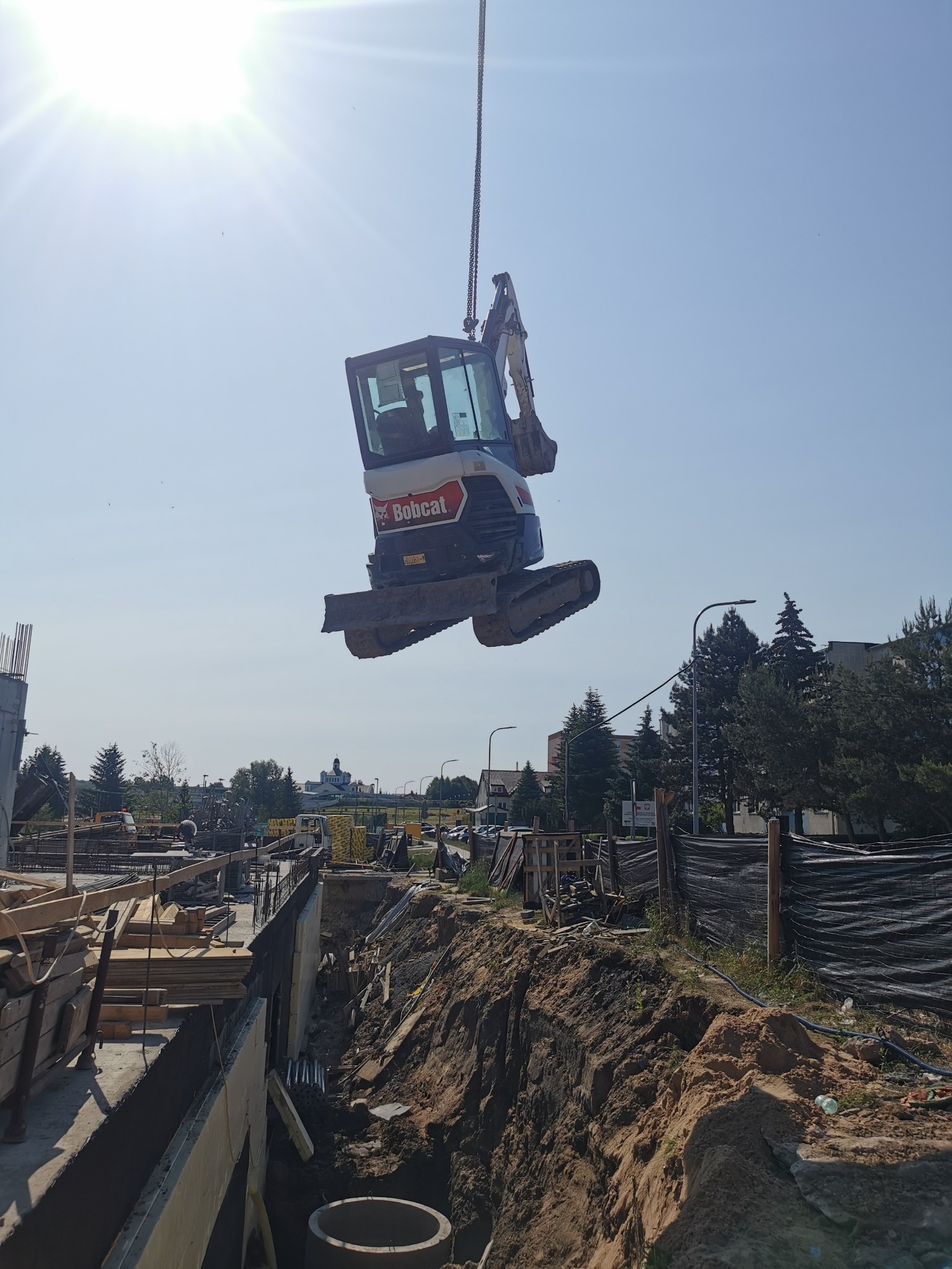 Mini koparka Bobcat podnoszona na placu budowy za pomocą dźwigu, widoczny wykop i elementy konstrukcyjne fundamentów. Słoneczny dzień, błękitne niebo.