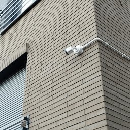 LANOPTIC - Kamera monitoringu zamontowana na rogu budynku z cegły, widoczne okno z żaluzjami. Białe rurki prowadzą kabel. Ujęcie z dołu.