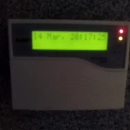 Panel alarmowy z wyświetlaczem LCD wskazującym datę i godzinę oraz diodami LED. Urządzenie w obudowie z tworzywa sztucznego.