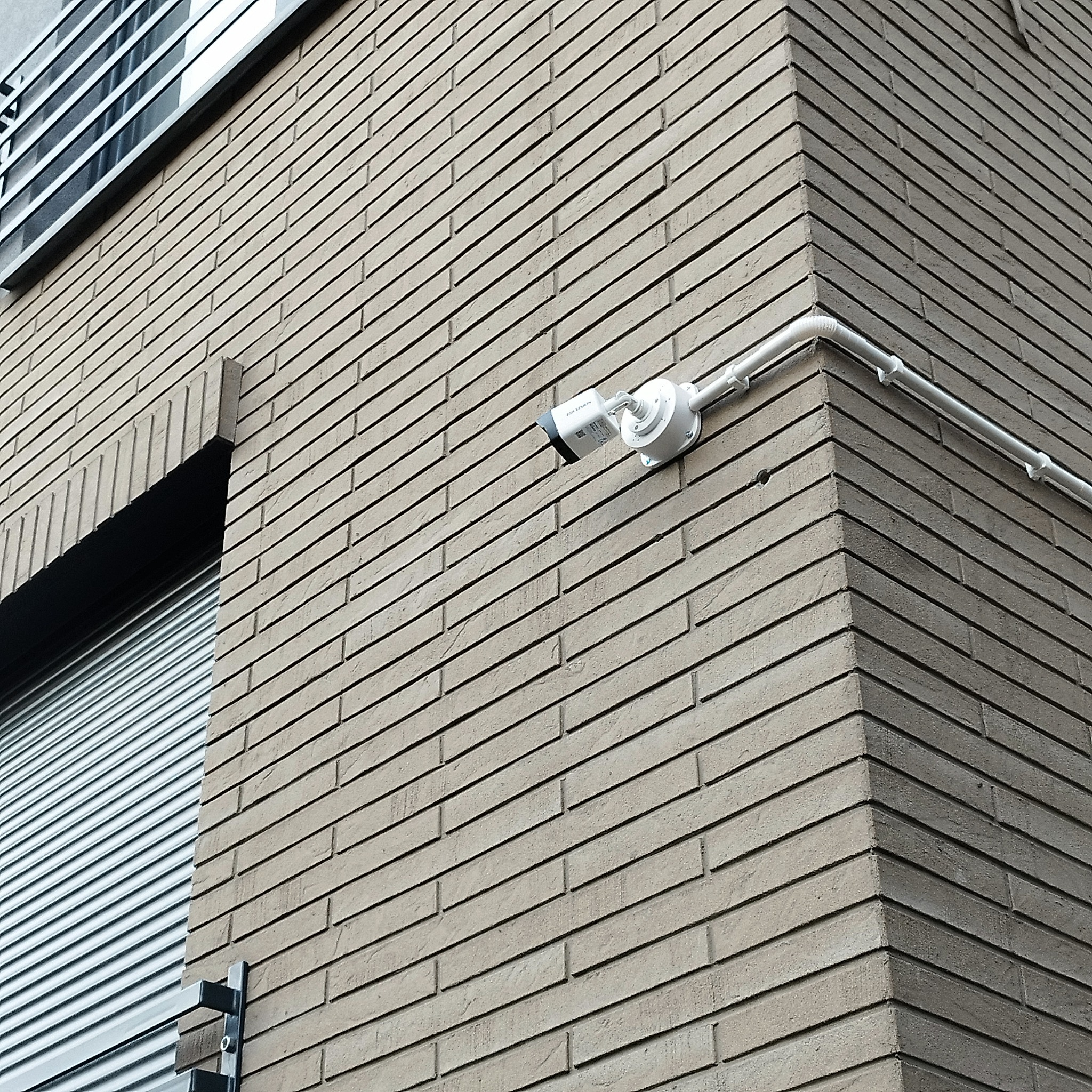 Kamera monitoringu zamontowana na rogu budynku z cegły, widoczne okno z żaluzjami. Białe rurki prowadzą kabel. Ujęcie z dołu.