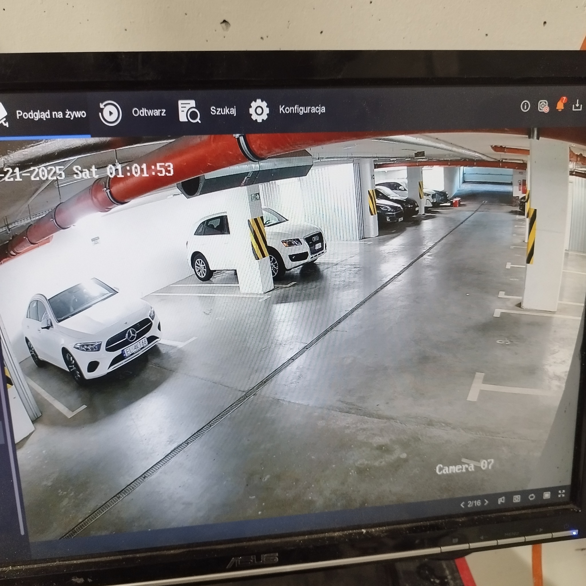 Obraz z kamery monitoringu w garażu podziemnym. Widoczne zaparkowane samochody, kolumny i rury pod sufitem. Na ekranie monitora widoczny numer kamery i data.
