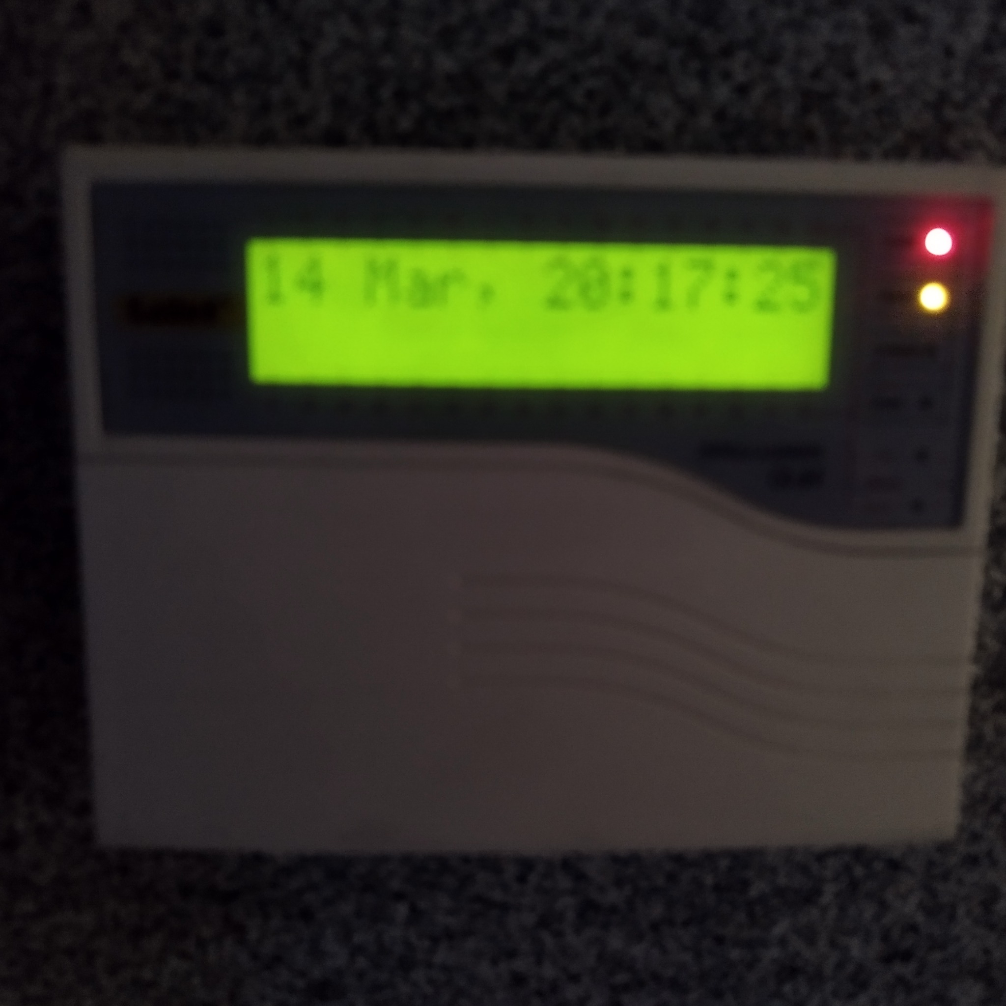 Panel alarmowy z wyświetlaczem LCD wskazującym datę i godzinę oraz diodami LED. Urządzenie w obudowie z tworzywa sztucznego.