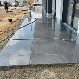 ANBI-BRUK Andrzej Białek - Układanie szarych płyt na tarasie. Widoczne krzyżyki dystansowe, przygotowane podłoże z kruszywa i odbicie nieba na mokrej powierzchni płyt.