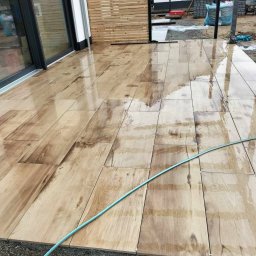 ANBI-BRUK Andrzej Białek - Ułożona, mokra, drewnopodobna kostka na tarasie przy domu. W tle widoczna drewniana ażurowa ścianka działowa i budynek w trakcie prac wykończeniowych.