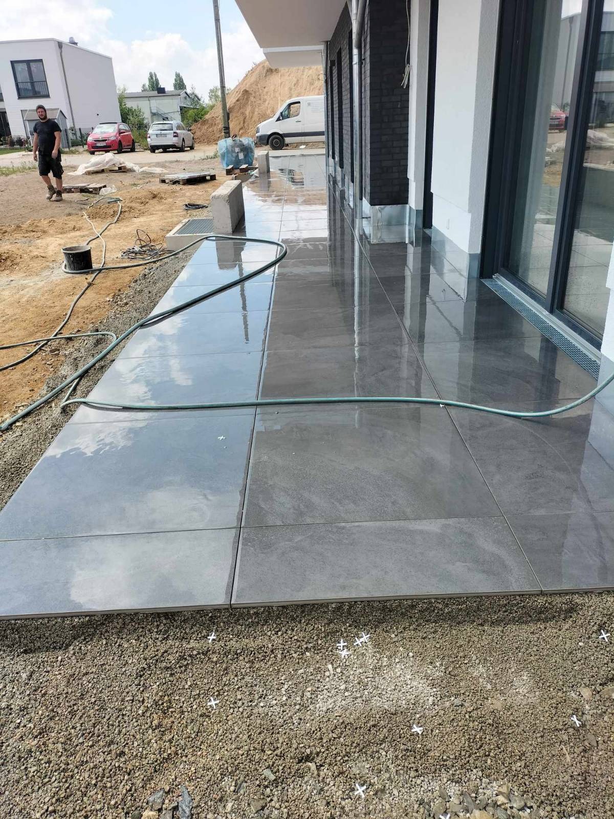 Układanie szarych płyt na tarasie. Widoczne krzyżyki dystansowe, przygotowane podłoże z kruszywa i odbicie nieba na mokrej powierzchni płyt.