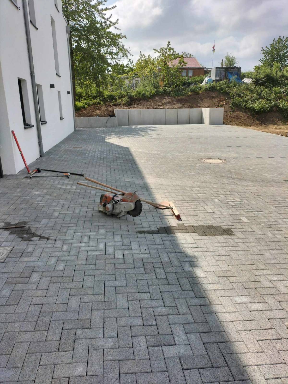 Świeżo ułożona, szara kostka brukowa w jodełkę na podjeździe, z widocznymi narzędziami brukarza i piłą do cięcia kostki. W tle biały budynek i zielona roślinność.