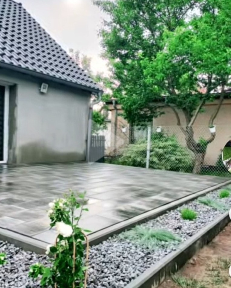 Nowoczesny taras z szarej kostki brukowej przy domu z ciemnym dachem, rabata z jasnymi kamykami i roślinami wzdłuż krawędzi tarasu, zielone drzewa w tle.