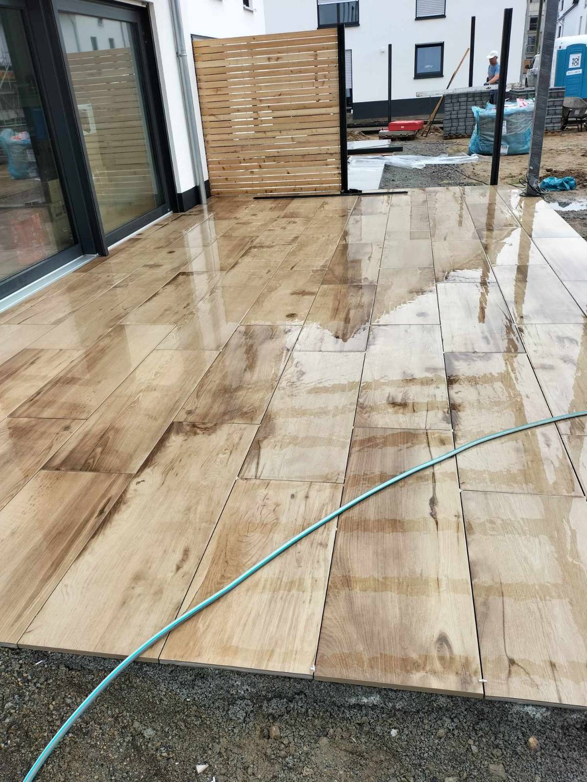 Ułożona, mokra, drewnopodobna kostka na tarasie przy domu. W tle widoczna drewniana ażurowa ścianka działowa i budynek w trakcie prac wykończeniowych.