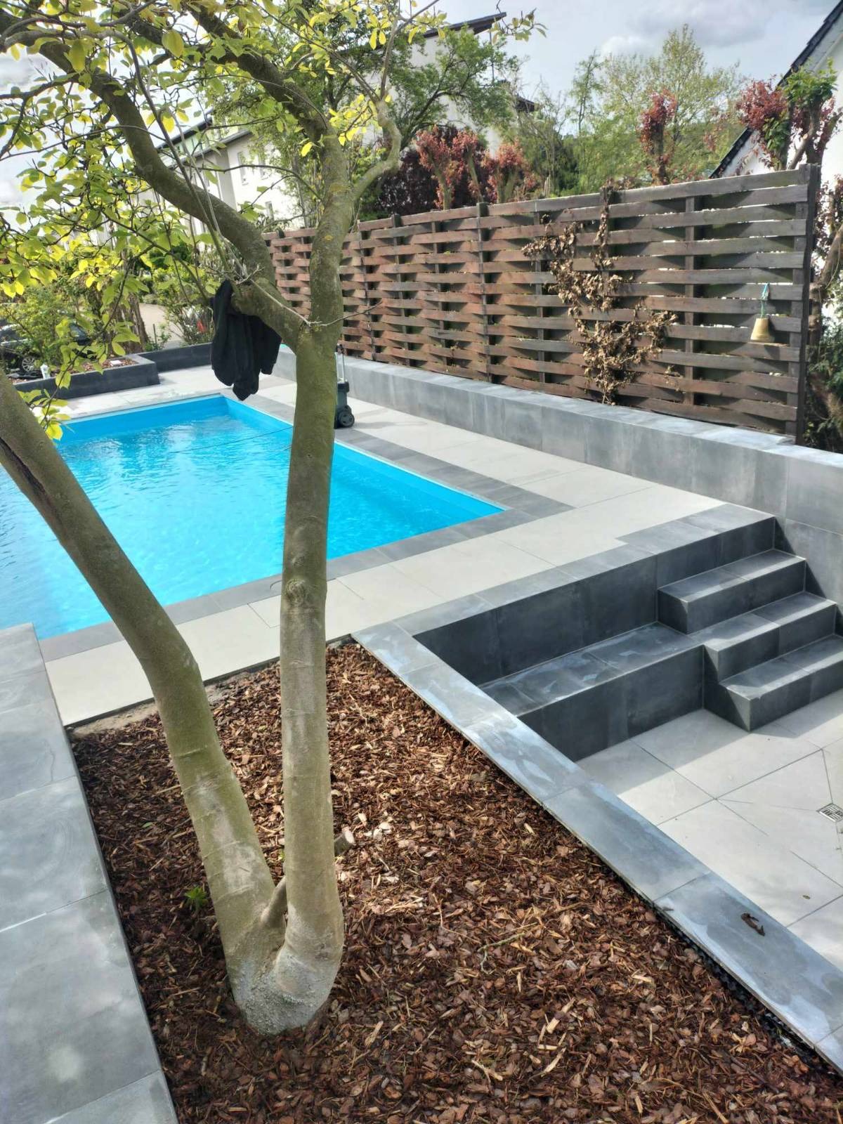 Nowoczesny ogród z basenem: szara kostka, drewniany ażurowy płot i ozdobna kora wokół drzewa. Schody z kostki prowadzą do basenu.