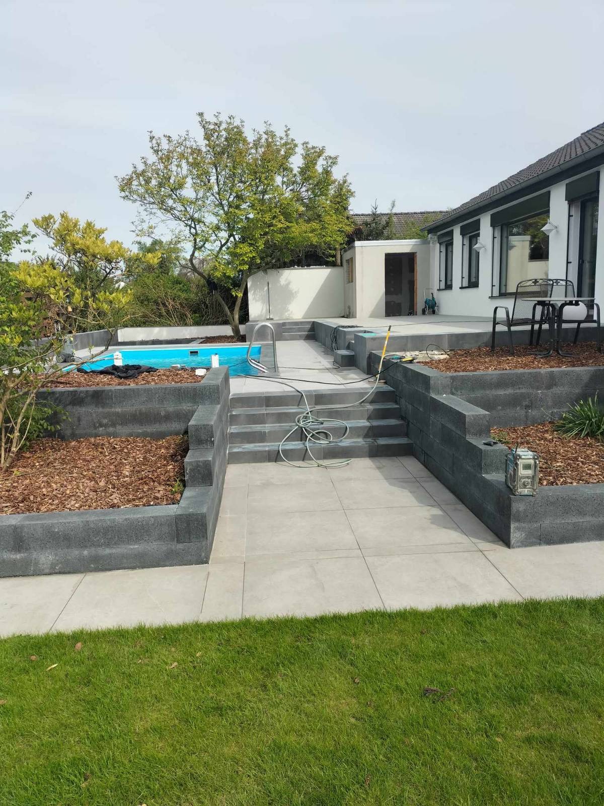 Nowoczesny taras z jasnych płyt, schody z kostki, obrzeża rabat z korą, basen w tle, zielony trawnik. Estetyczne zagospodarowanie przestrzeni przydomowej.