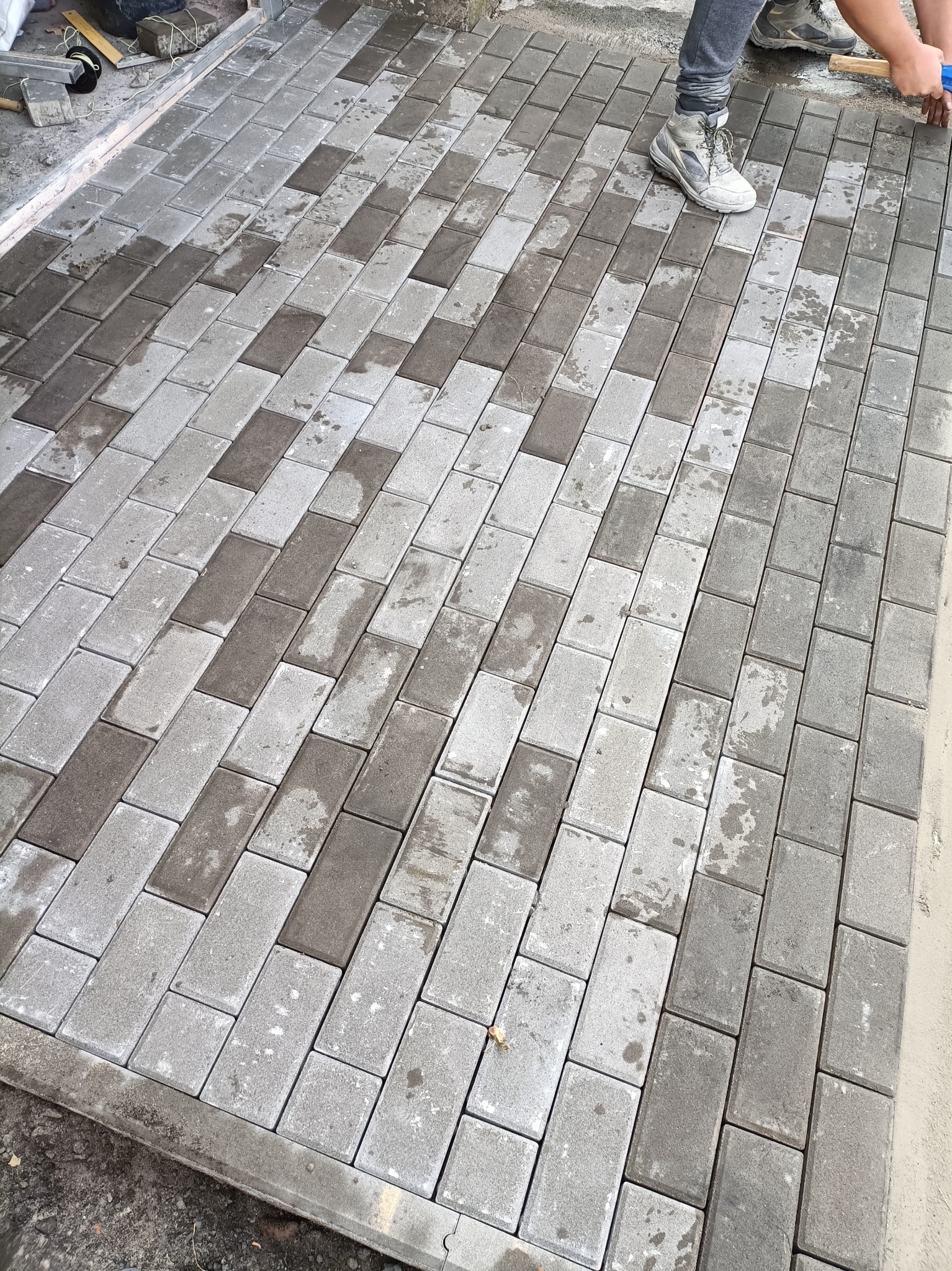 Świeżo ułożona, dwukolorowa kostka brukowa w odcieniach szarości, częściowo mokra, z widocznymi narzędziami i fragmentem buta osoby wykonującej prace.