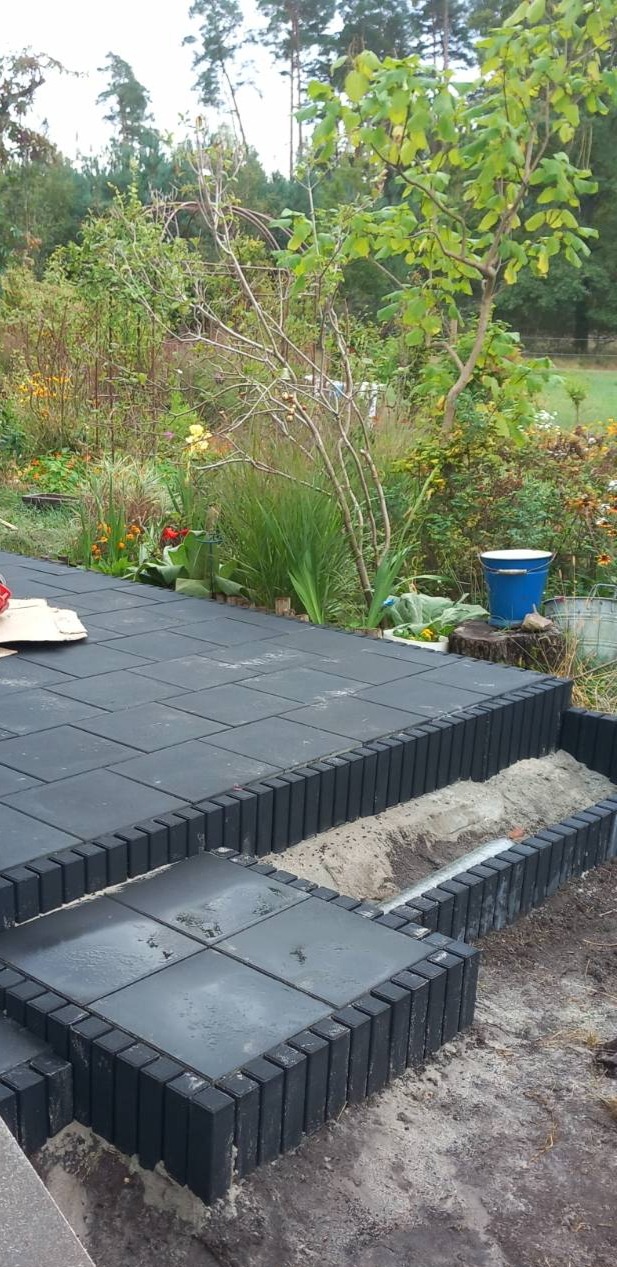 Układanie czarnej kostki brukowej w ogrodzie. Widoczne fragmenty ułożonej nawierzchni i przygotowane podłoże z obrzeżami. W tle roślinność ogrodowa.