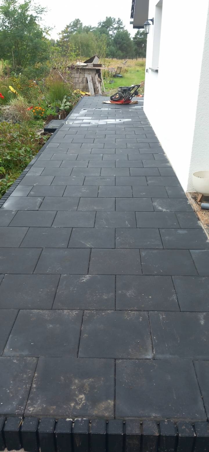 Ułożona, grafitowa kostka brukowa tworząca ścieżkę obok domu. W tle widoczna zieleń i maszyna do ubijania kostki. Ujęcie z perspektywy chodnika.