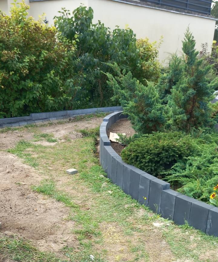 Obrzeże z szarej kostki betonowej oddziela rabatę z zielenią od ścieżki z piaskiem i trawą. Częściowo ułożone, w tle roślinność i budynek.