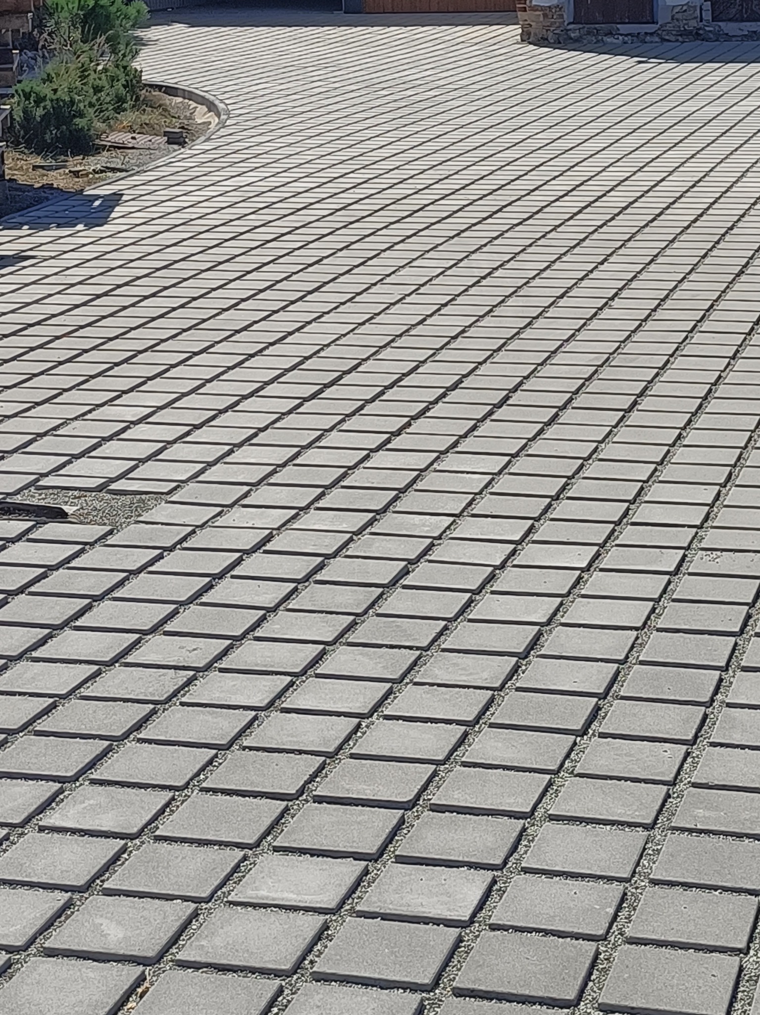 Szeroki kadr na ułożoną, szarą kostkę brukową w kwadratowym wzorze, z drobnym żwirem między kostkami. Fragment uszkodzonej nawierzchni w lewym dolnym rogu.