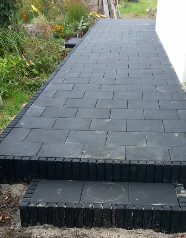 Ułożona, ciemna kostka brukowa tworząca ścieżkę z niskimi schodami, obrzeża z pionowo ułożonych kostek. Widok na ogród w tle, przy ścianie budynku.