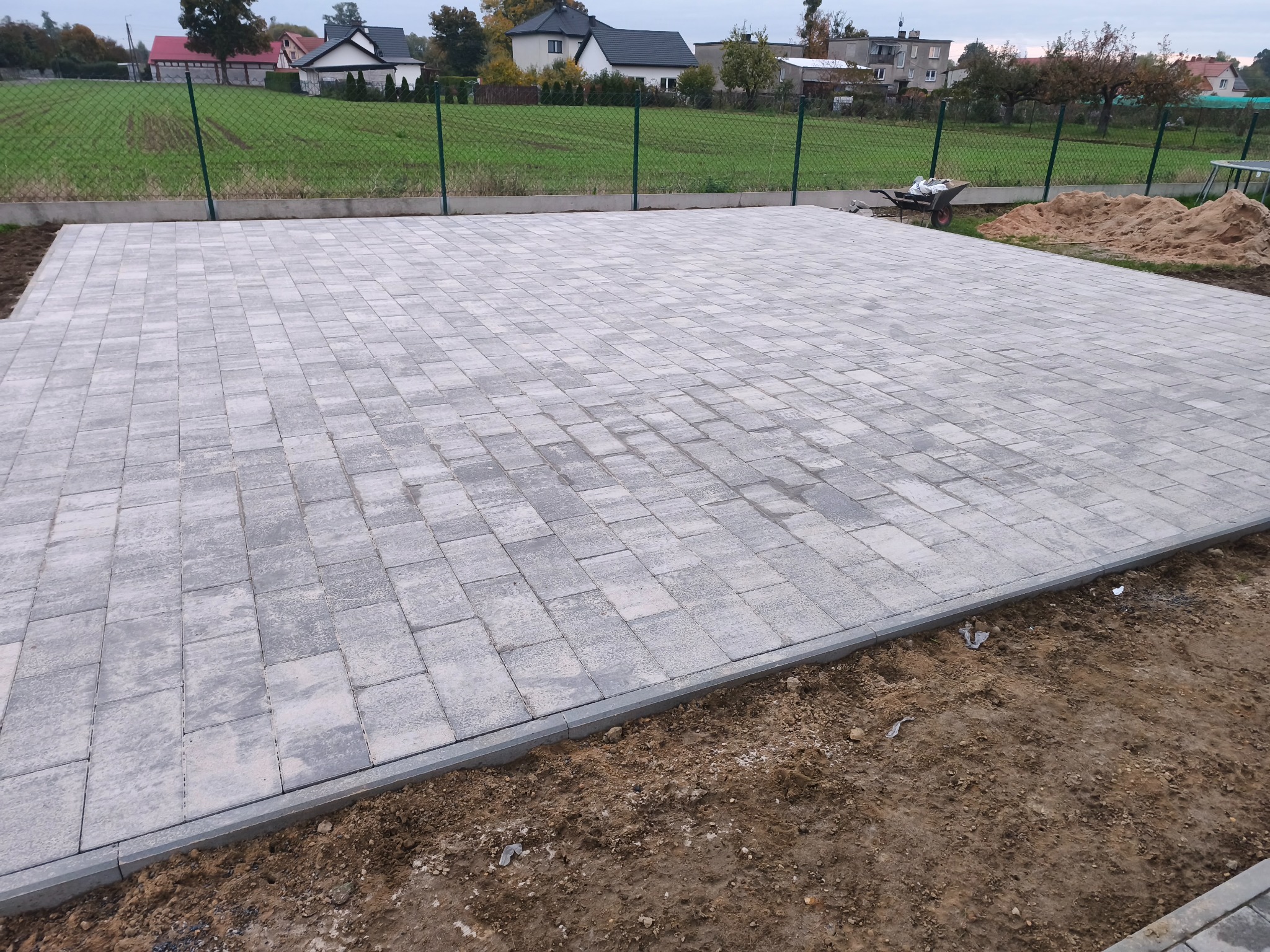 Ułożona, szara kostka brukowa na posesji z widocznym ogrodzeniem i domami w tle. Uwagę zwraca równa powierzchnia i estetyczne wykończenie obrzeżami.