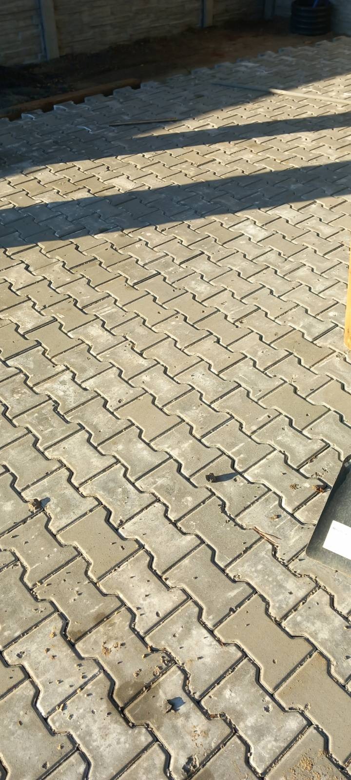 Ułożona, szara kostka brukowa typu 'I' na posesji, widoczna z góry, z fragmentem ogrodzenia w tle. Częściowe zacienienie podkreśla fakturę.